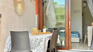 Ein Essbereich mit einem Tisch für zwei Personen, einem Korbstuhl und Blick durch eine Glastür in ein Schlafzimmer.