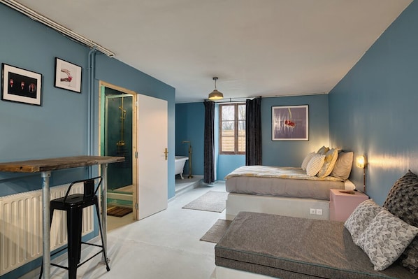 A bedroom with a bed, a desk, and a chair. There is a bathroom visible through an open door.