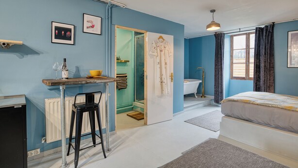 A bedroom with a bed, a desk with a chair, a radiator, a window with curtains, and a door leading to a bathroom. - Suite Privée, Bain, Kitchenette, en Champagne. (Orges)