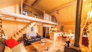 A log cabin interior with a wooden staircase, a living area with a sofa and coffee table, a kitchen area, and a wood-burning stove. - Uniquely landscaped log cabin with spa! (Lac-aux-Sables)