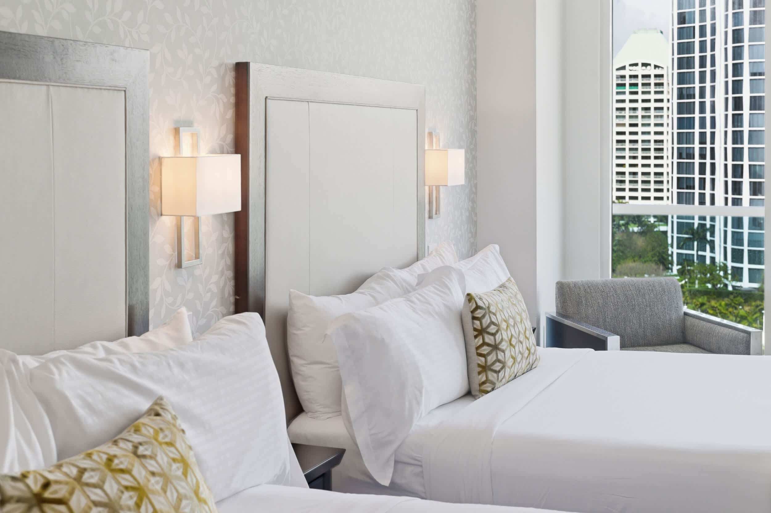 Ein Hotelzimmer mit einem großen Bett, zwei Nachttischen, einem Sessel und einem Fenster mit Blick auf die Stadt.