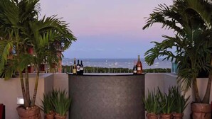Zona de bar con vista al mar y barcos, rodeada de plantas en macetas.