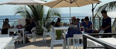 Un área de comedor al aire libre con sillas y mesas de plástico blanco, sombrillas grandes y vista al mar.