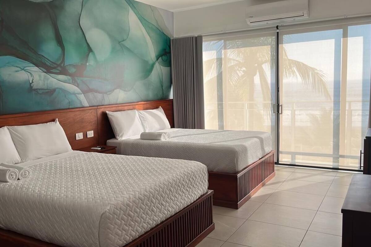 A hotel room with two beds, a large abstract mural on the wall, a sliding glass door to the balcony, and a dark wooden desk.