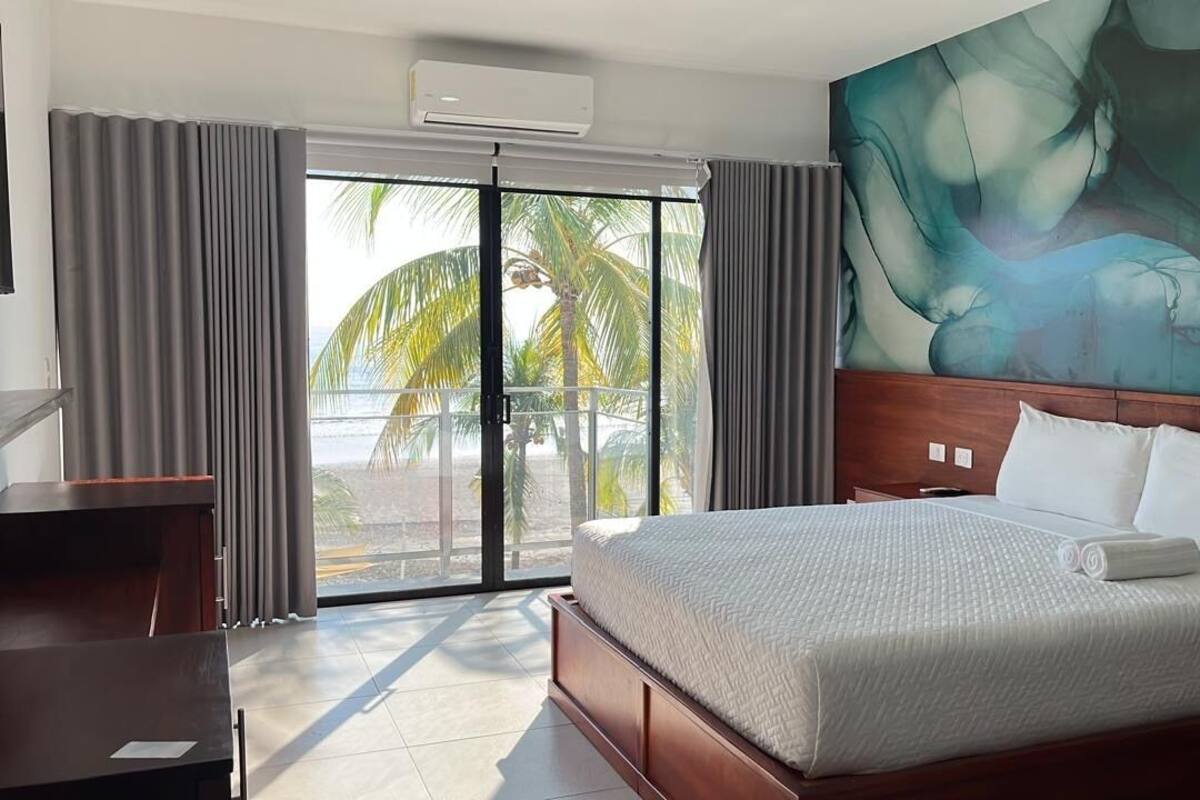 A hotel room with a large bed, a desk, and a view of palm trees through a sliding glass door.
