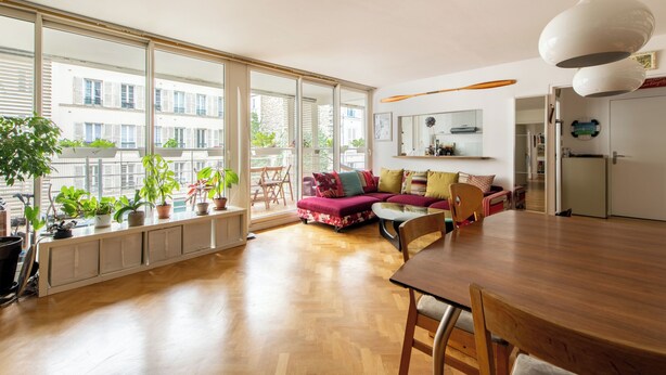 Een ruime woonkamer met een houten vloer, een groot raam, een eettafel en een bank met kleurrijke kussens.