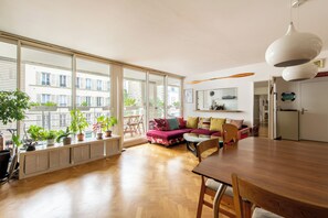 A spacious living area with a wooden floor, a large window, a dining table, and a sofa with colorful cushions. - Quiet family apartment (110 m²) with a terrace, near Montmartre (Paris)