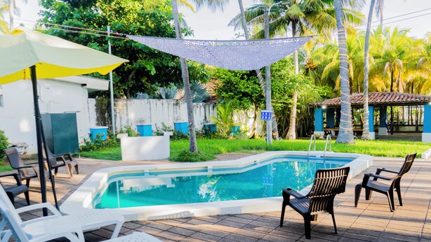 Una piscina con sillas de descanso y una zona sombreada, rodeada de vegetación y un pequeño edificio.