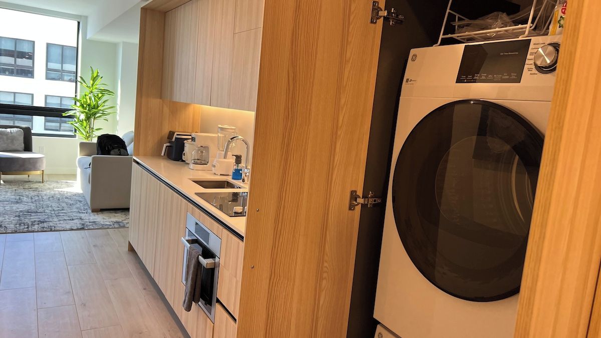 A modern kitchen with wooden cabinets, a built-in washing machine, and a dining area with a grey sofa.