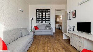 Een moderne woonkamer met een grijze bank, rode en zwarte kussens, een salontafel en een flatscreen televisie op een witte kast.
