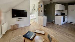 En moderne lejlighed med et fjernsyn ophængt på væggen, et køkken med hvide skabe og et spisebord med en bog og en skål.