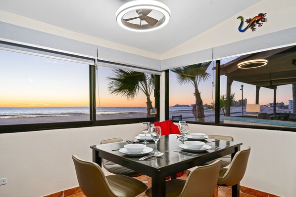 A dining area with a table set for four, large windows offering a view of the beach and ocean at sunset. - Las Gaviotas 413 (Puerto Peñasco)