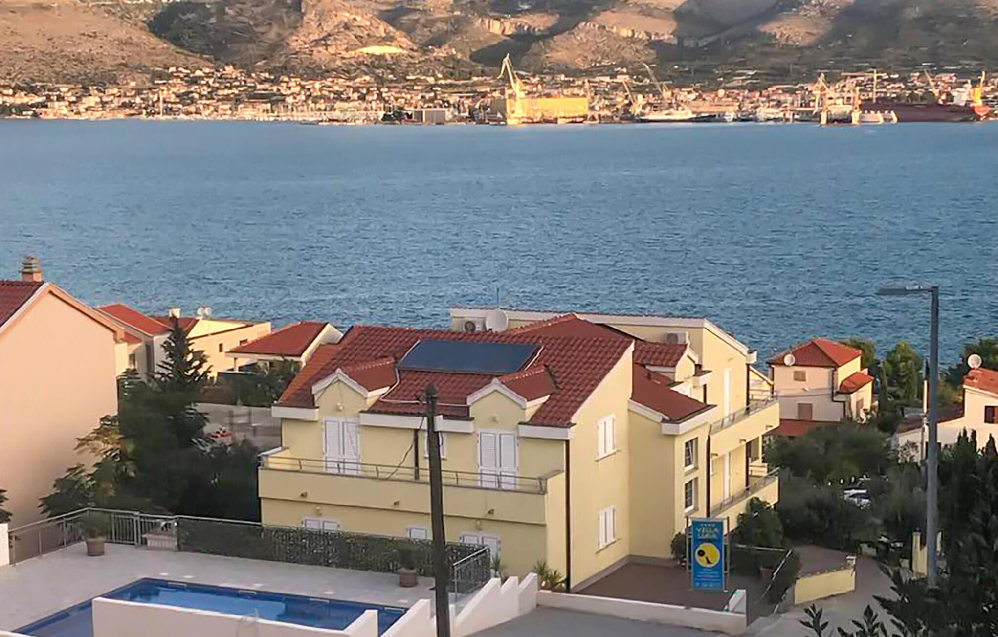 En kystbygning med swimmingpool, røde tegltage og udsigt til havet og en by i det fjerne.
