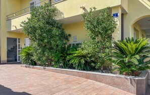 Ein Gebäude mit gelber Fassade, einer Treppe, die zu einem Eingang führt, und einer mit Pflanzen bepflanzten, gefliesten Terrasse.