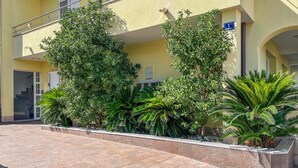 Un bâtiment à la façade jaune, avec un escalier menant à l’entrée et une terrasse carrelée agrémentée de différentes plantes.