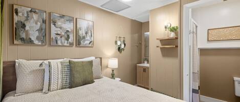 A bedroom with a large bed, a nightstand, a lamp, a wall-mounted shelf with a plant, and abstract artwork.
