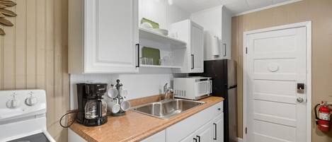 A compact kitchen with white cabinets, a stainless steel sink, a coffee maker, a microwave, and a black refrigerator.