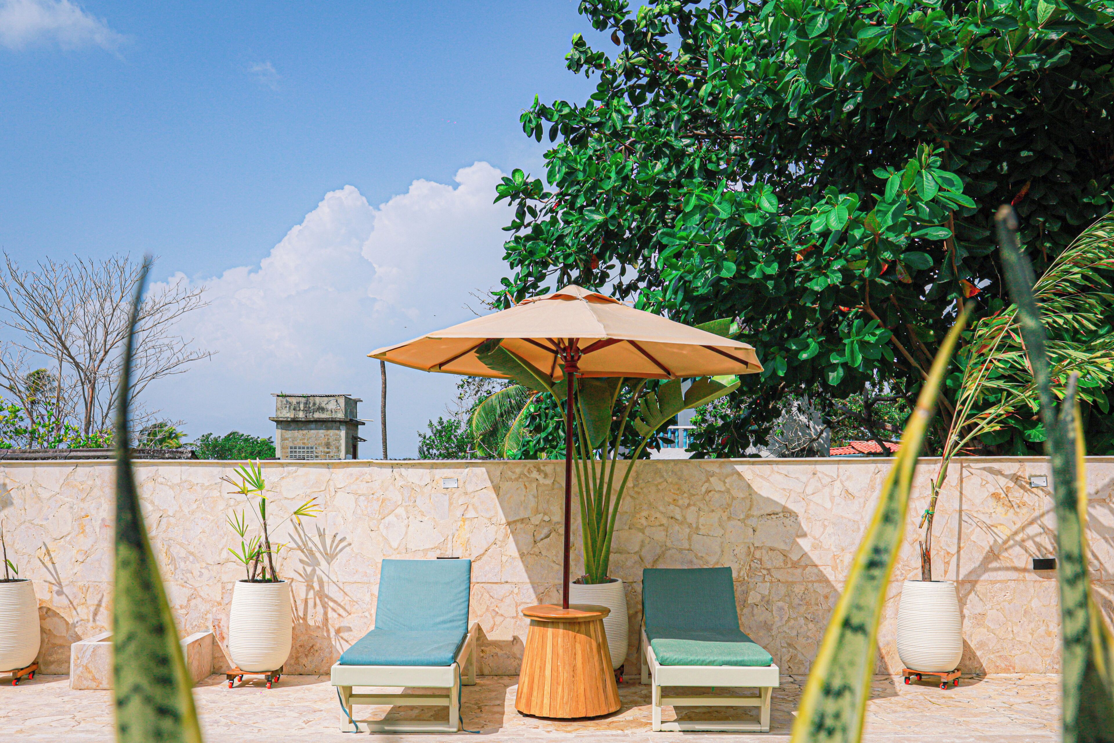 Two lounge chairs, a wooden table, and an umbrella in a patio area with a stone wall and potted plants.