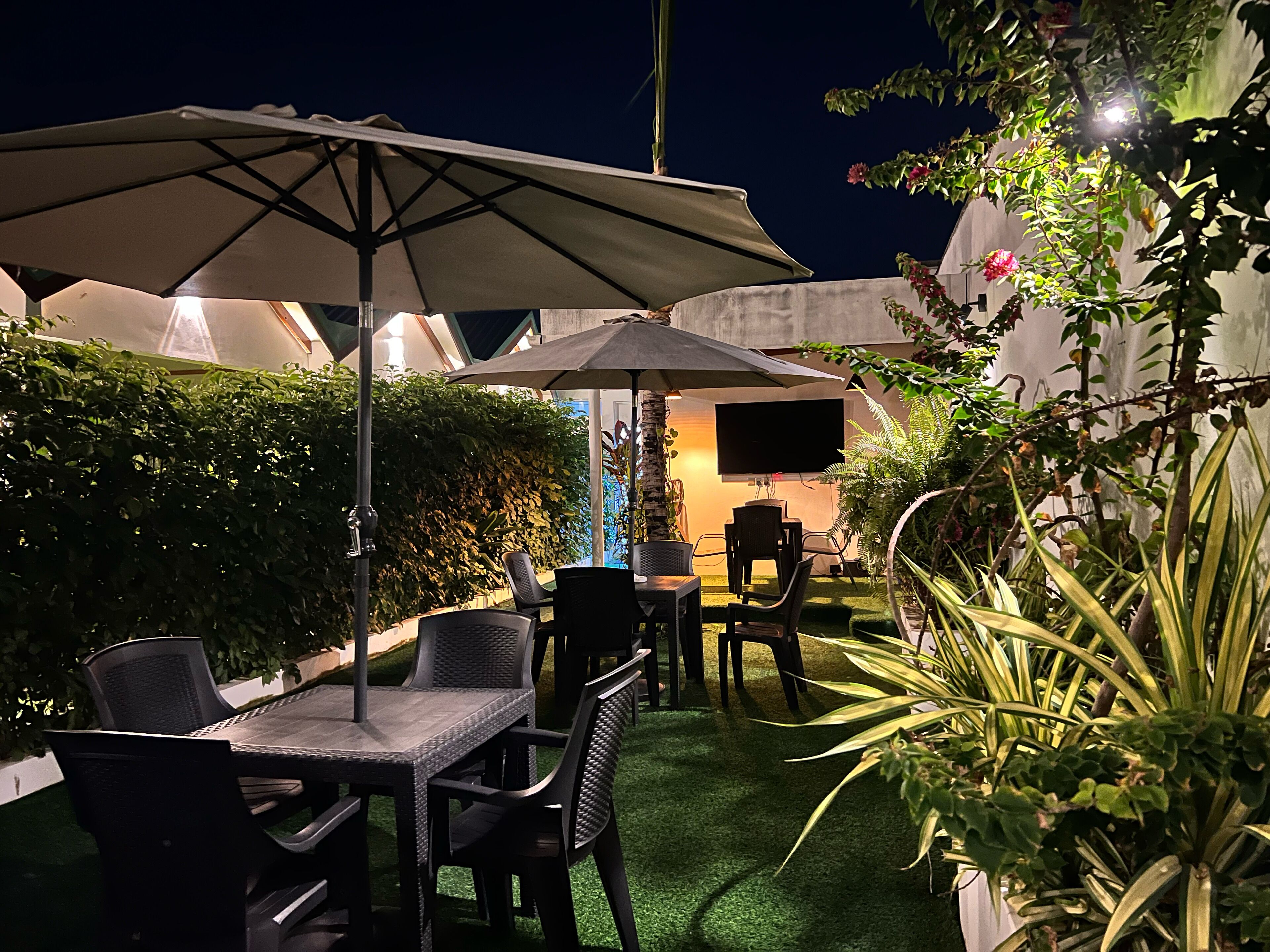 Area patio esterna con tavoli e sedie sotto ombrelloni, immersa nel verde e con uno schermo televisivo.