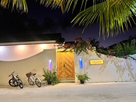 Entrada de um edifício com porta de madeira, duas bicicletas estacionadas e uma placa com a inscrição 'HANDHUVARU MALDIVES'.