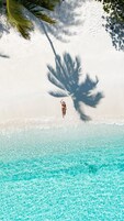 Uma pessoa em pé numa praia com águas turquesa cristalinas e uma palmeira projetando uma sombra.
