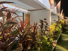 Interior de hotel moderno com um corredor longo, ladeado por plantas em vasos, teto de madeira e uma janela com cortinas.