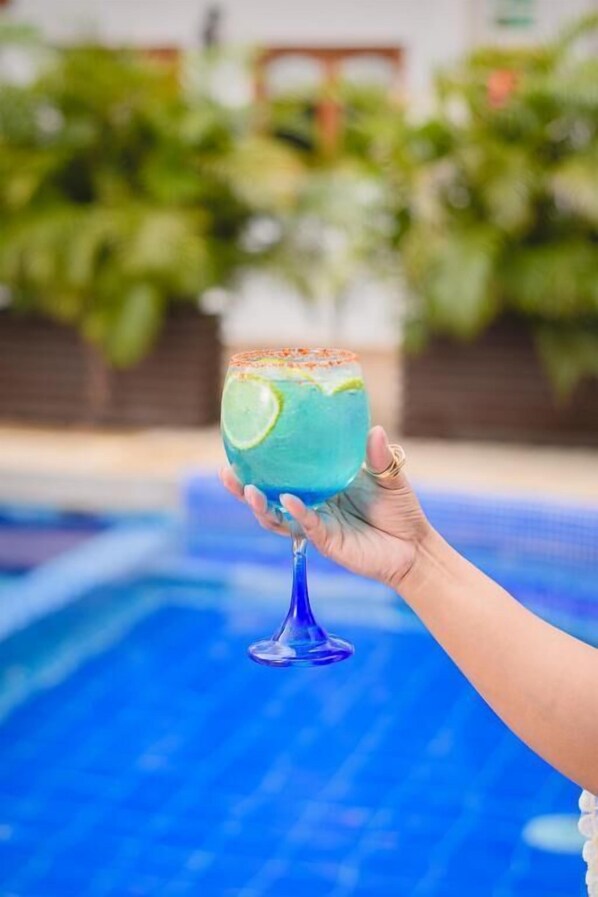 A hand holding a blue cocktail glass with a lime wedge, near a swimming pool. - Hotel Abitti (Taganga)
