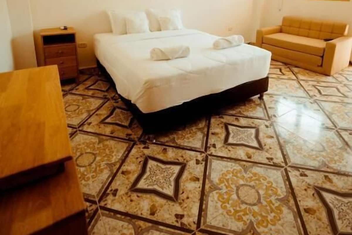 A hotel room with a bed, a wooden nightstand, a sofa, and patterned floor tiles.