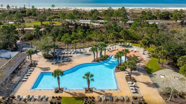 Ein Luftbild eines Resorts mit einem zentral gelegenen Schwimmbad, umgeben von Liegestühlen und Palmen, mit einem Strand und dem Meer im Hintergrund.
