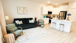 Ein modernes Wohnzimmer mit einer Couch, Sesseln, einem Couchtisch und einer Küchenzeile mit Kühlschrank, Mikrowelle und Barhocker.
