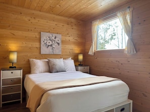 Un dormitorio con paneles de madera, una cama, dos mesitas de noche, un revistero y una ventana con cortinas, además de un cuadro en la pared.