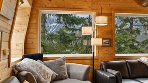 En mysig stuga med träpanel på väggarna, ett stort fönster med utsikt över träd och berg, en soffa med kuddar och en golvlampa.