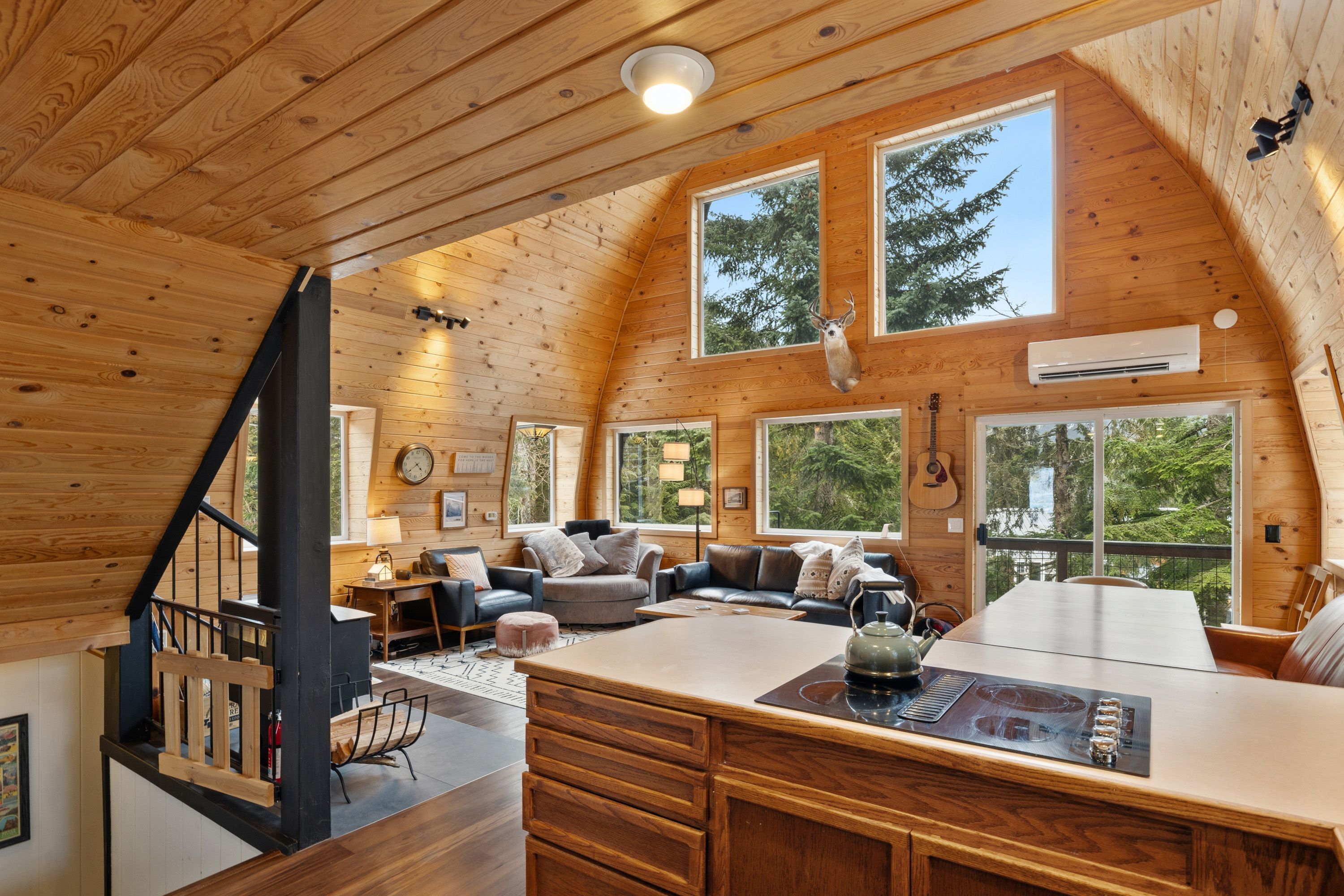 A spacious cabin with a wooden interior, large windows, a central living area with a sofa and coffee table, and a staircase.