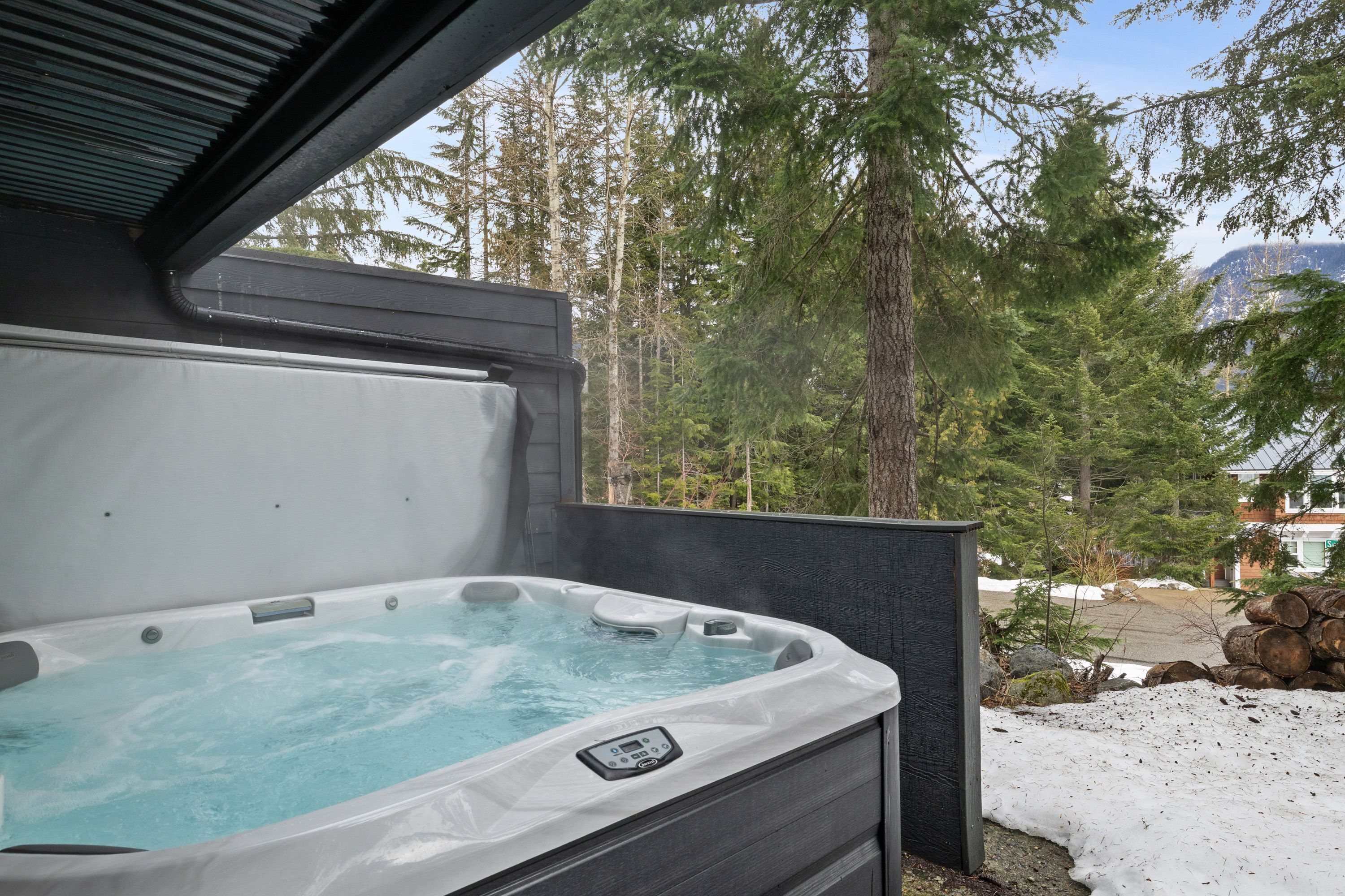 Un bain à remous doté d’un panneau de commande, entouré d’une terrasse en bois et surplombant un paysage enneigé avec des arbres et des montagnes en arrière-plan.