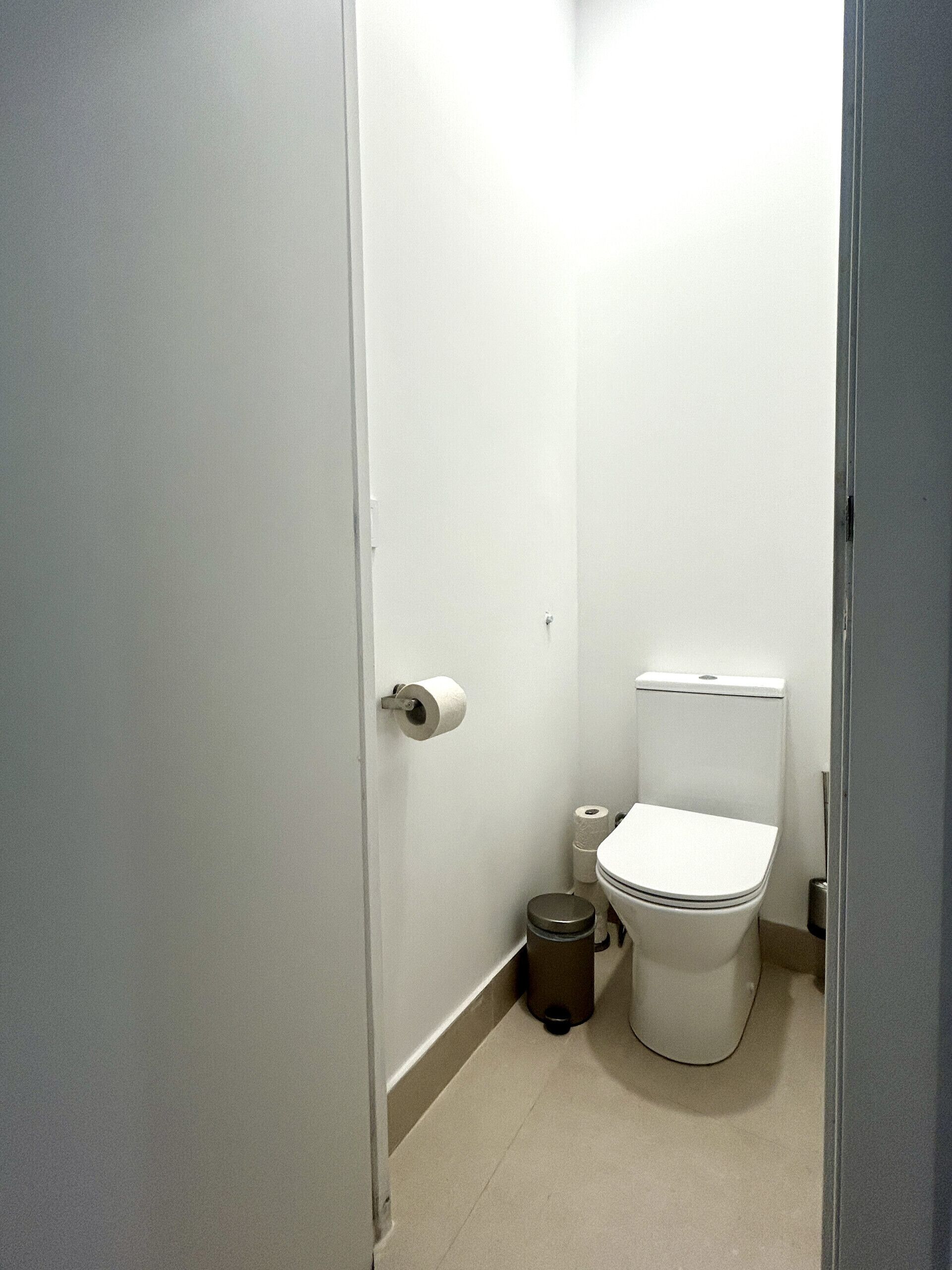 A small, narrow bathroom with a toilet, a trash can, and a roll of toilet paper on the wall.