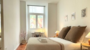 Een slaapkamer met een bed, een bureau, een stoel en een raam met uitzicht op kleurrijke gebouwen.