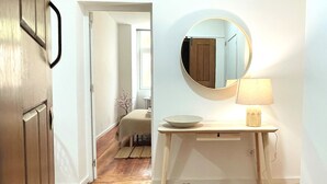 Een hal met een houten vloer die leidt naar een badkamer, met een houten deur, een ronde spiegel, een nachtkastje met een lamp en een kleine plant.