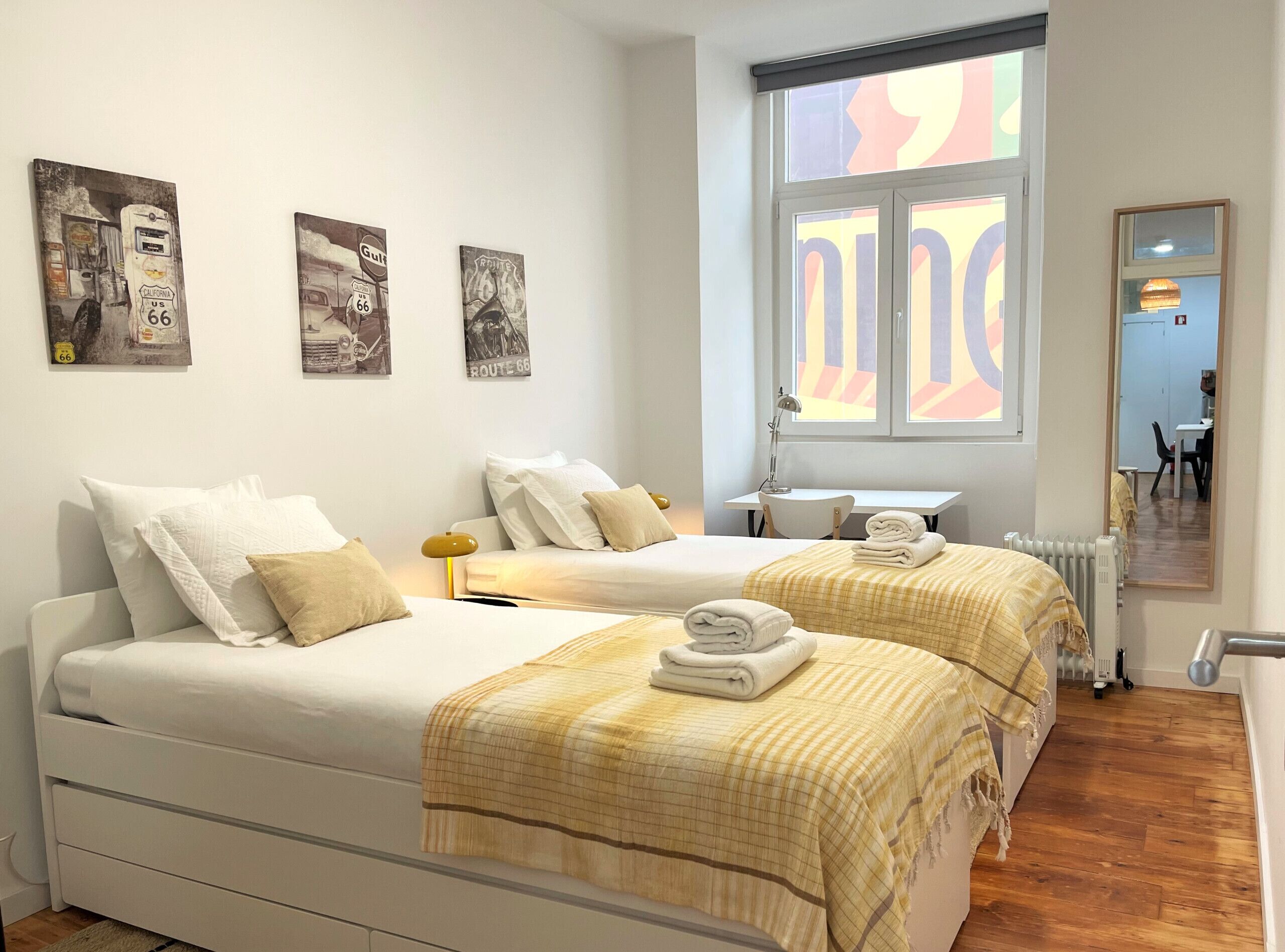 A hotel room with two beds, a desk, a chair, a window with a view, and artwork on the wall.