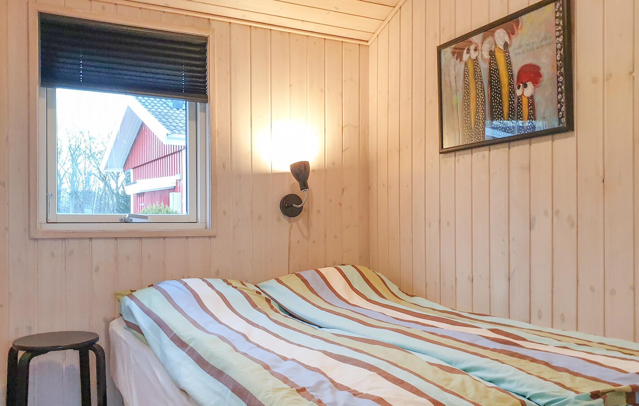 Una habitación con cama, ventana con persianas, luz en la pared y un cuadro.