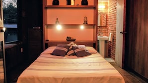 A bedroom with a bed, pillows, a shelf with decorative items, a clock, and a brick wall.