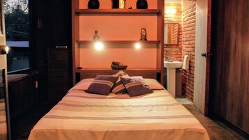 Une chambre à coucher avec un lit, des oreillers, une étagère avec des objets décoratifs, une horloge et un mur en briques.