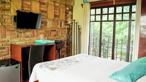 A room with a brick wall, a television mounted on the wall, a bed with a patterned bedspread, and a window with a view of greenery.