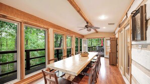 Una larga mesa de comedor con sillas, ventanas amplias y un ventilador de techo en una habitación con vigas de madera y vistas a árboles.