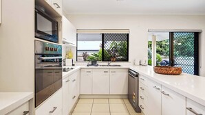 A modern kitchen with white cabinetry, stainless steel appliances, a large window with a view of greenery, and a tiled floor. - Family Entertainer in Kanimbla - Space & Privacy (Kanimbla)