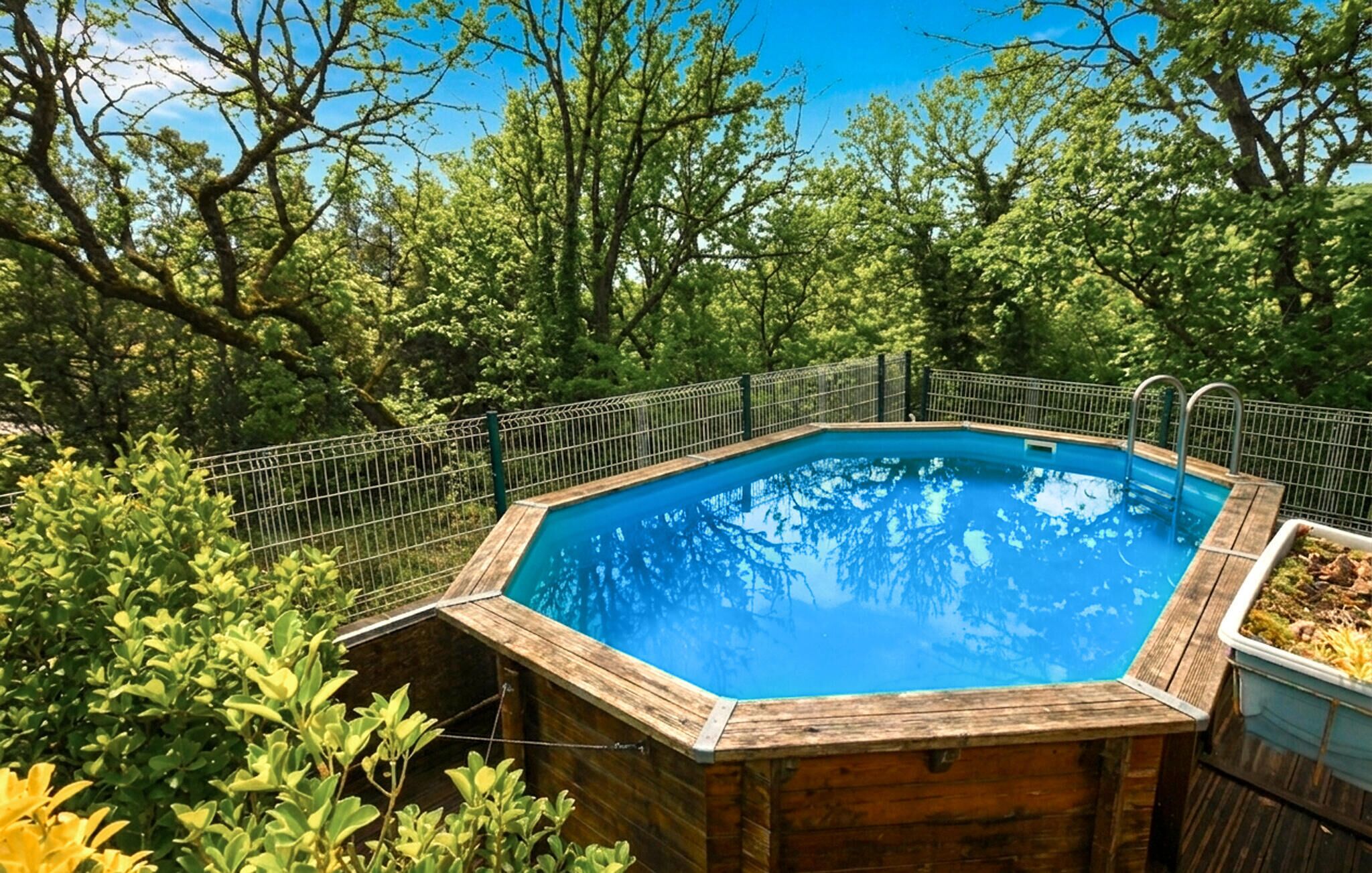 Une piscine hexagonale en bois, entourée d’une clôture métallique, située au cœur d’une forêt luxuriante et verdoyante.
