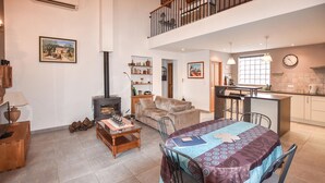 A spacious living area with a high ceiling, a fireplace, a dining table with chairs, a sofa, and a kitchen area with a window and a clock. - Maison avec piscine 1 (Mormoiron)