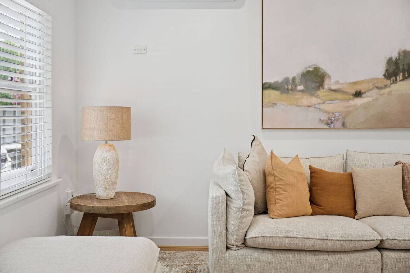 A living room with a beige sofa, a round wooden side table with a lamp, and a painting on the wall.