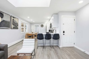 Eine moderne Küche mit weißen Schränken, ein Essbereich mit Holztisch und -stühlen sowie ein Wohnbereich mit Sofa und Couchtisch.