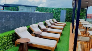 A rooftop lounge area with sun loungers and tables, surrounded by a green artificial turf and a decorative wall. - JJ JIVANA BALI (Tibubeneng)