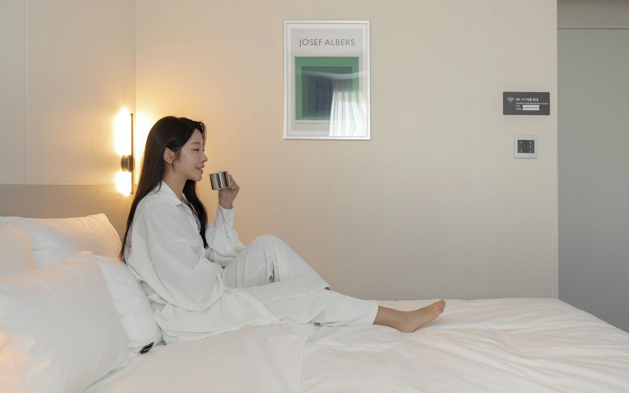 A person sitting on a bed in a hotel room with a framed artwork on the wall and a Wi-Fi sign.